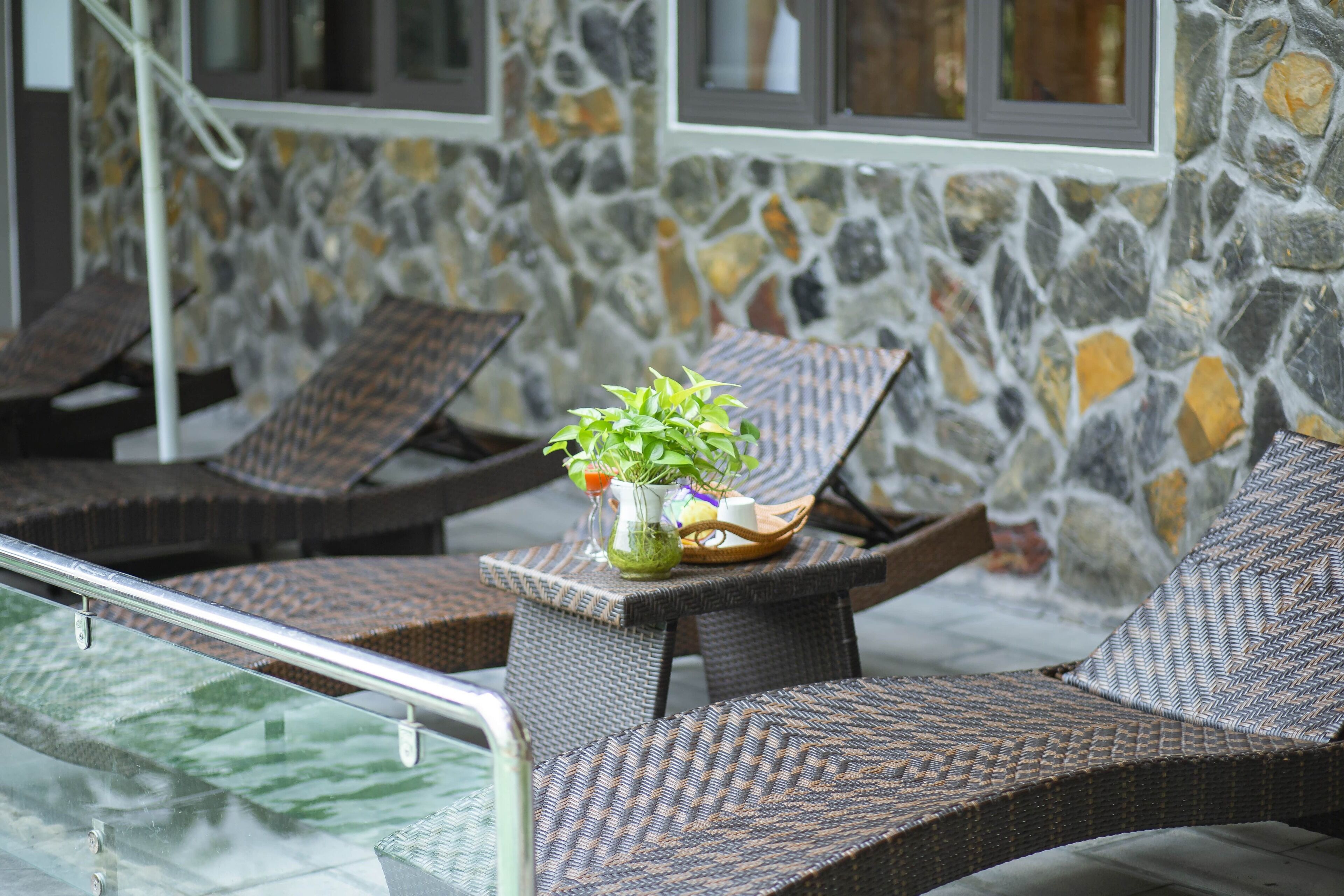 A poolside area with wicker lounge chairs, a small table with a plant, and a stone wall backdrop.