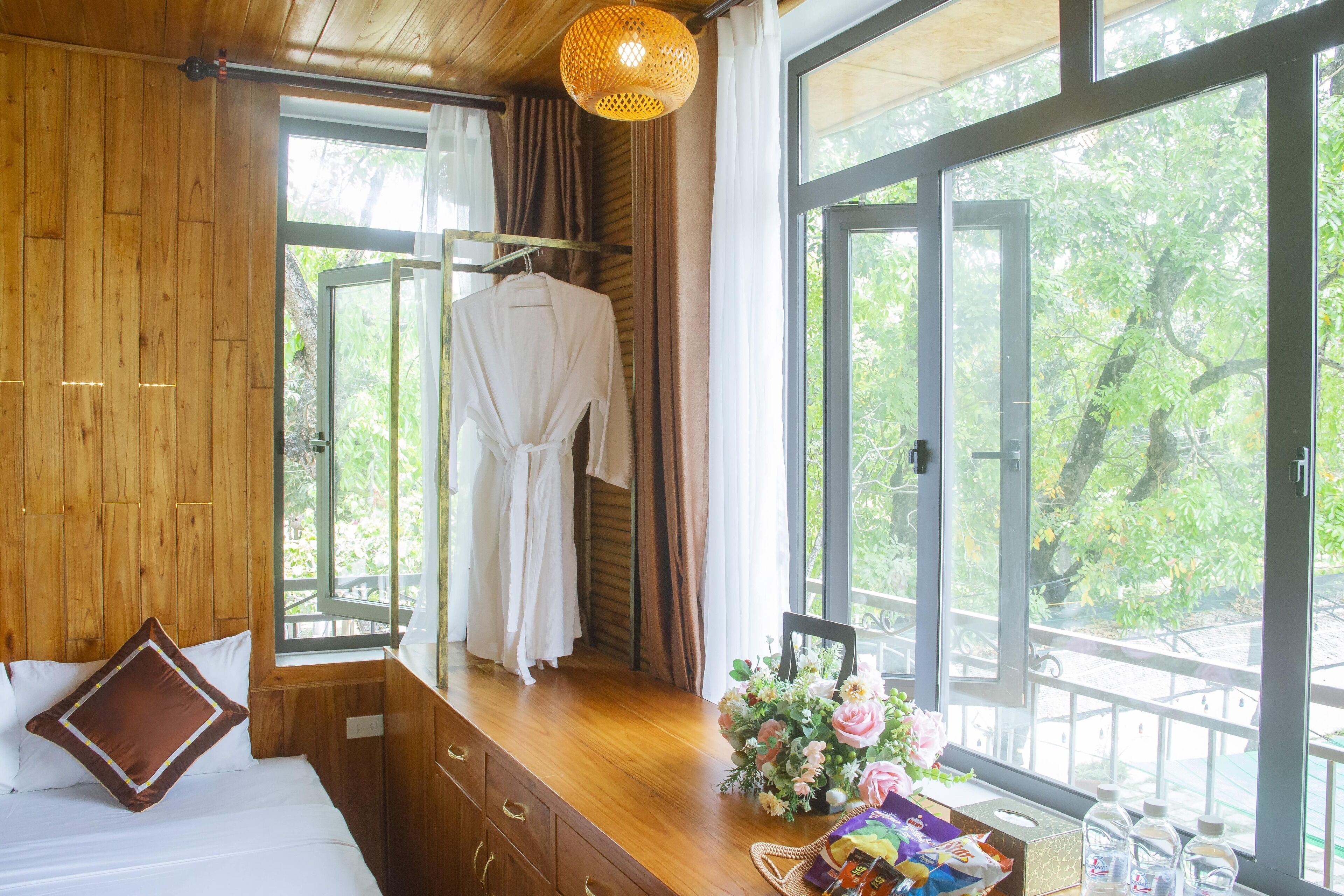 A wooden room with a bed, a window with a view of trees, a hanging robe, and a table with a vase of flowers and bottles.