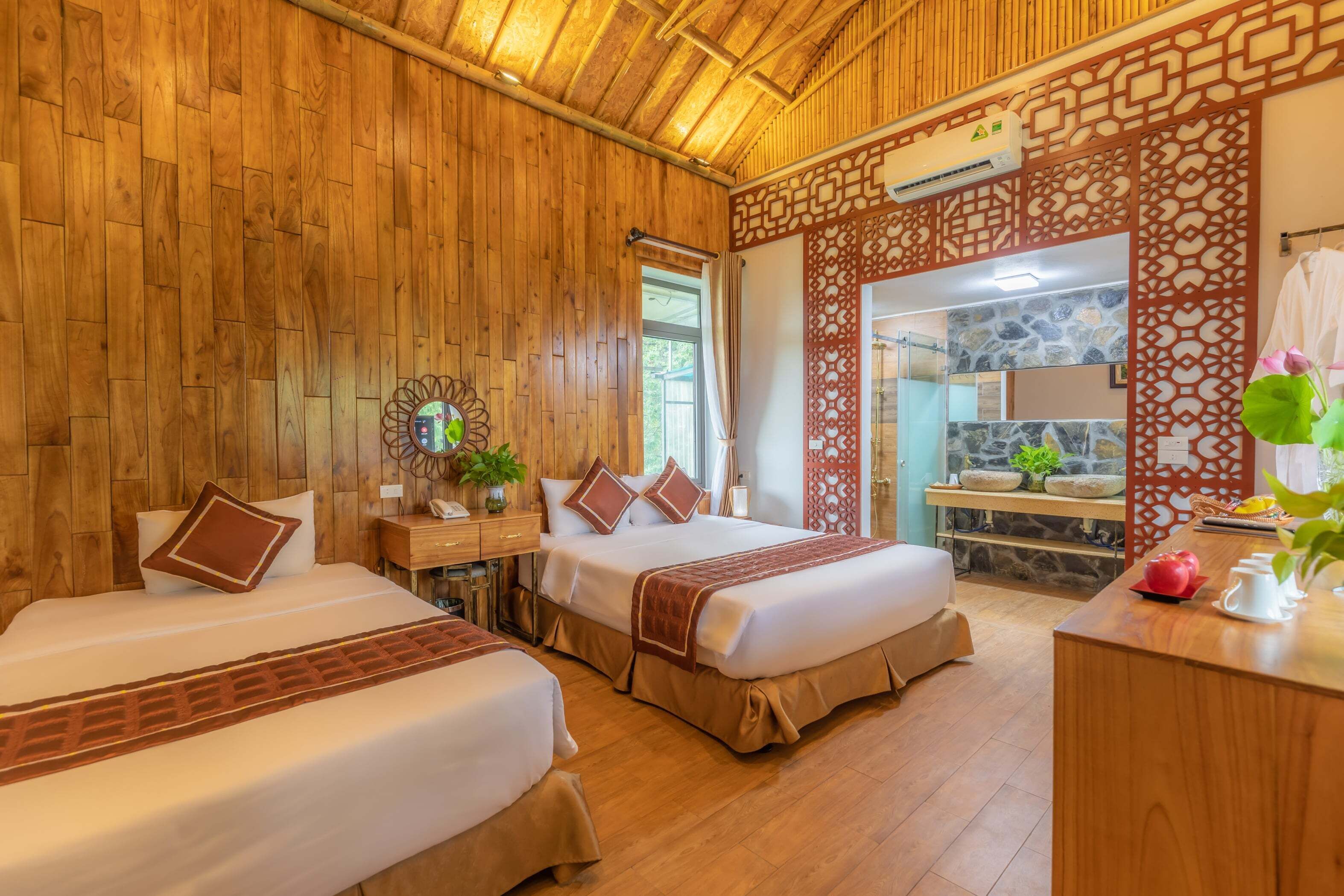 A wooden-themed room with two beds, a small desk, and a bathroom visible through a glass door.