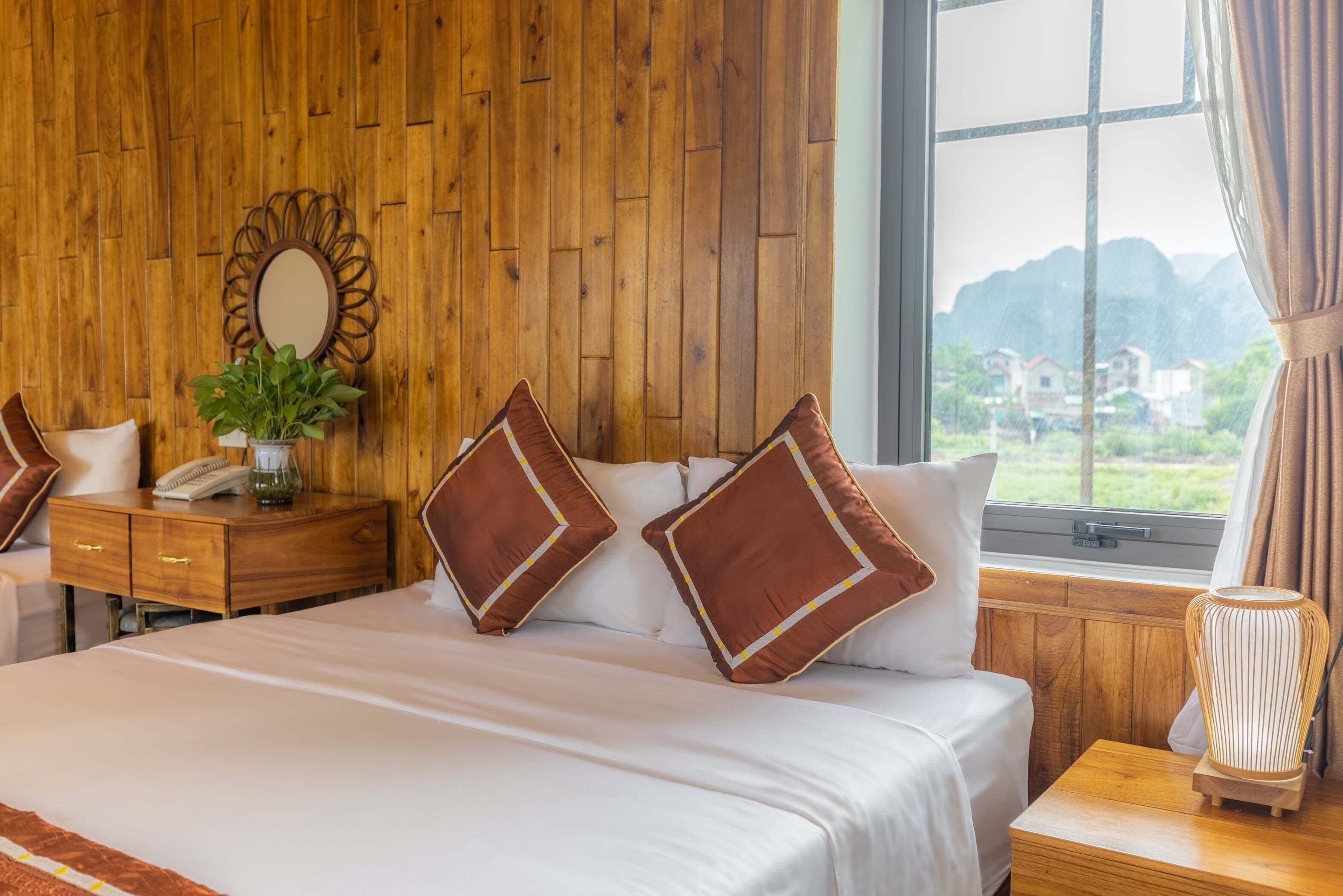 Ein Hotelzimmer mit einer Holzwand, einem Bett mit weißer Bettwäsche und braunen Kissen, einem Nachttisch mit Lampe sowie Blick auf Grünflächen und Gebäude durch ein Fenster.