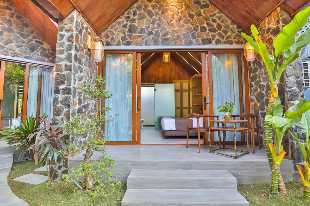 Une façade en pierre avec un toit en bois, un patio couvert avec un canapé et une table, et un escalier menant à l’intérieur.