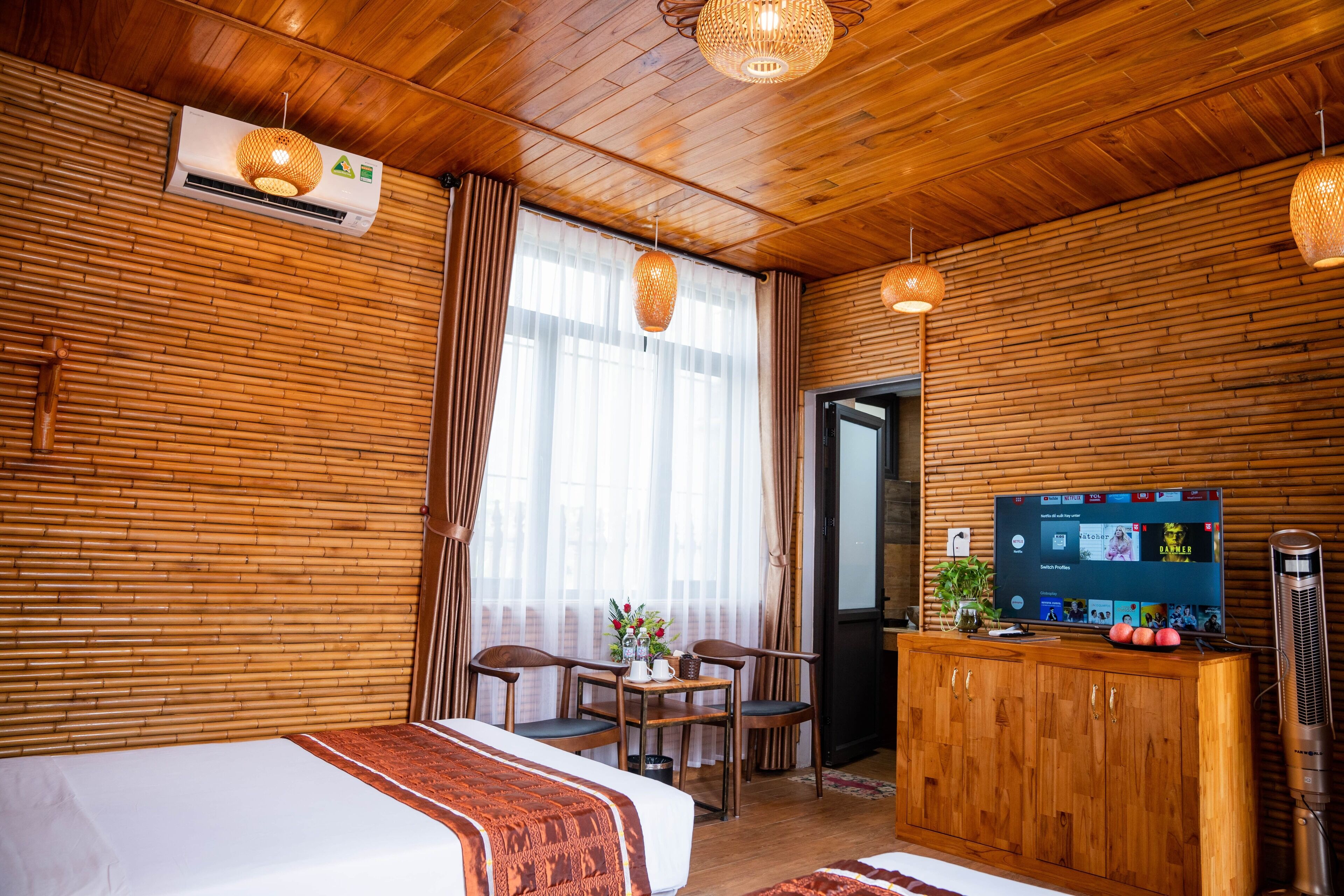 A hotel room with a wooden ceiling, a bed with a patterned blanket, a TV on a wooden cabinet, and a window with sheer curtains.