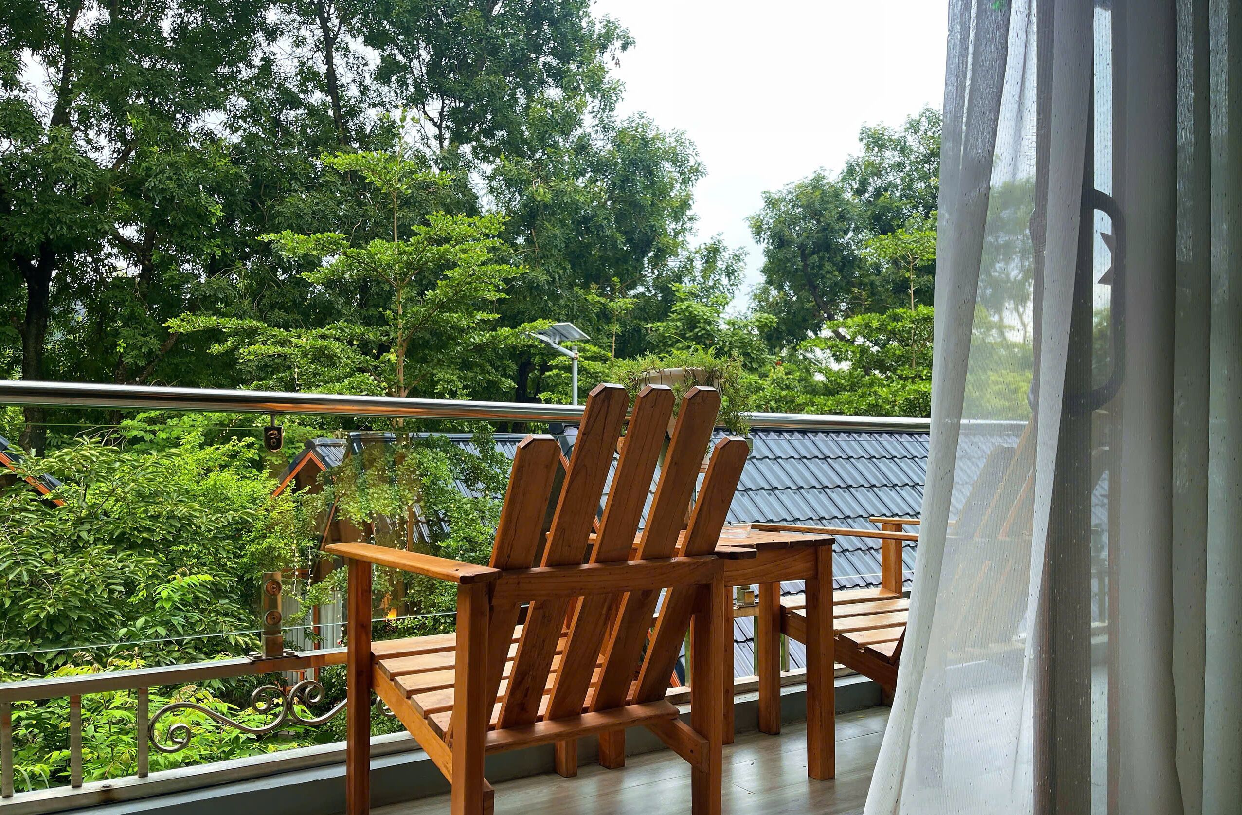 A balcony with wooden chairs and a table, overlooking a lush green landscape with trees and a metal roof.