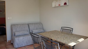 Um quarto pequeno com um sofá cinzento, uma mesa de metal e duas cadeiras. Há uma estante branca com objetos decorativos na parede.
