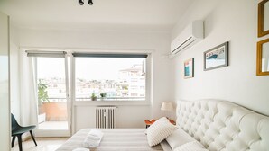 A hotel room with a large bed, a view of the city through a window, a radiator, and a small table with a lamp. - Bright studio in Trastevere/ ponte Testaccio (Roma)
