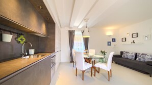 A modern kitchen with a central island, dining area with white chairs, and a sofa with patterned cushions. - BASTINICA KRK Superior Ap 3, OldTown, CityCenter (Krk)