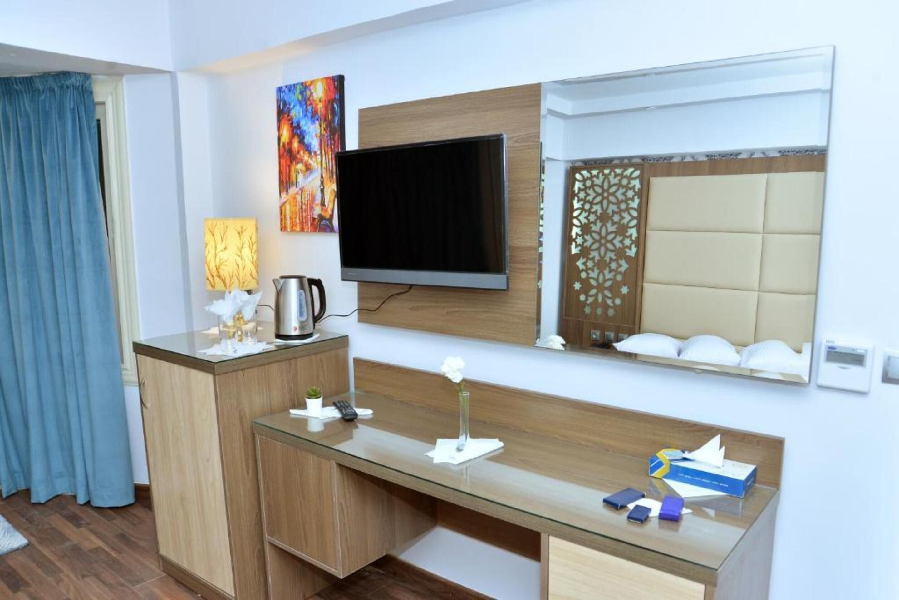 A hotel room with a wooden desk, a flat-screen TV, a decorative mirror, and a window with a patterned blind.