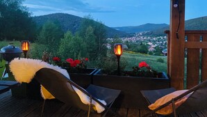 Eine Holzterrasse mit zwei Stühlen und einem Tisch, mit Blick auf ein Dorf und Hügel.