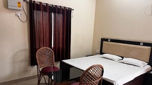 A hotel room with a single bed, two wicker chairs, a small table, and a window with curtains. - Sewadham Training Centre (Phulpur)