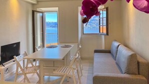 A room with a dining table and chairs, a sofa, a TV, and a view of a lake and mountains through the windows. - Casa Spasù: charming corner with a view 5 minutes from Cannobio, Lake Maggiore (San Bartolomeo)