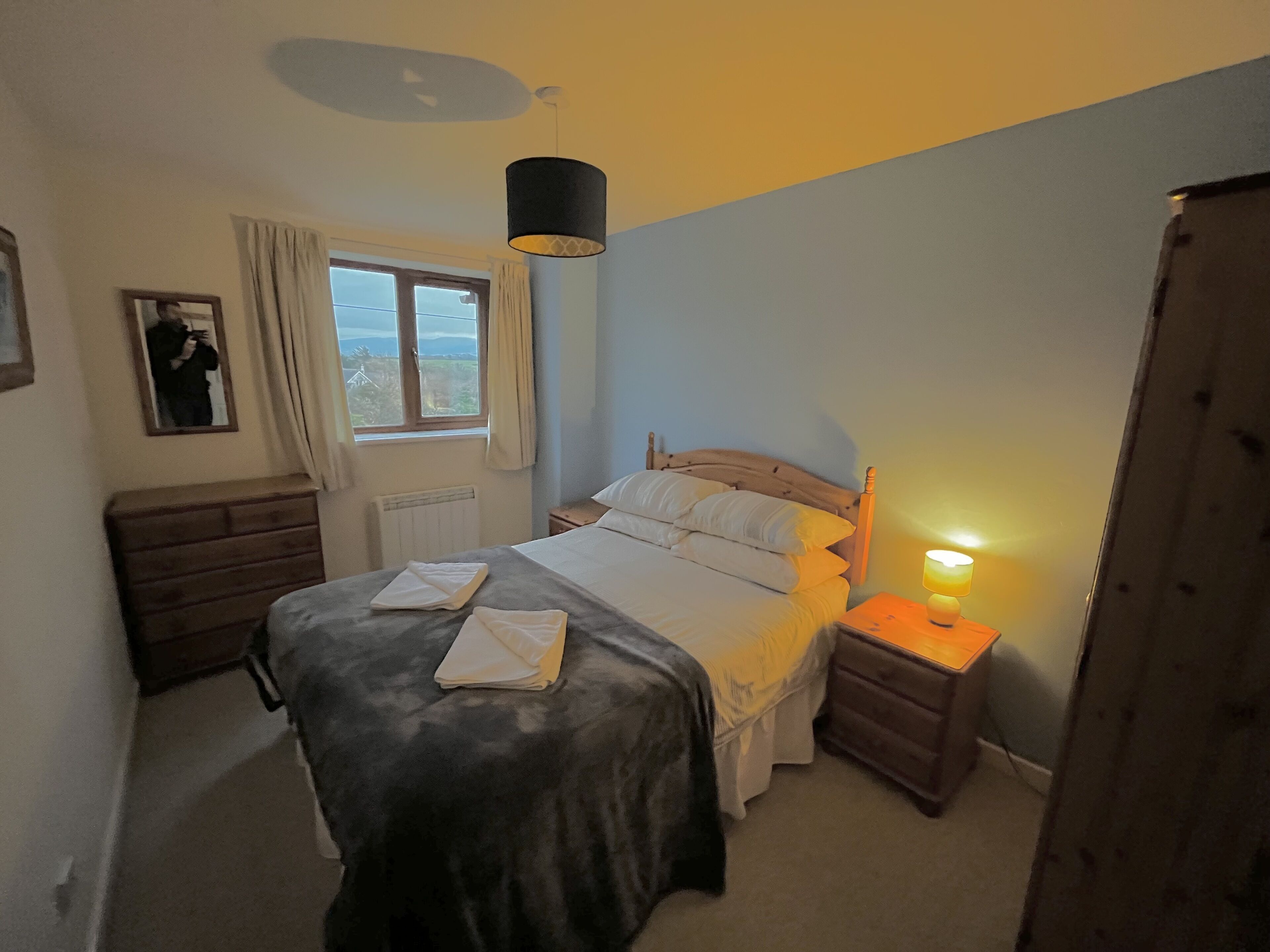 A bedroom with a bed, bedside table, lamp, window, and a mirror.