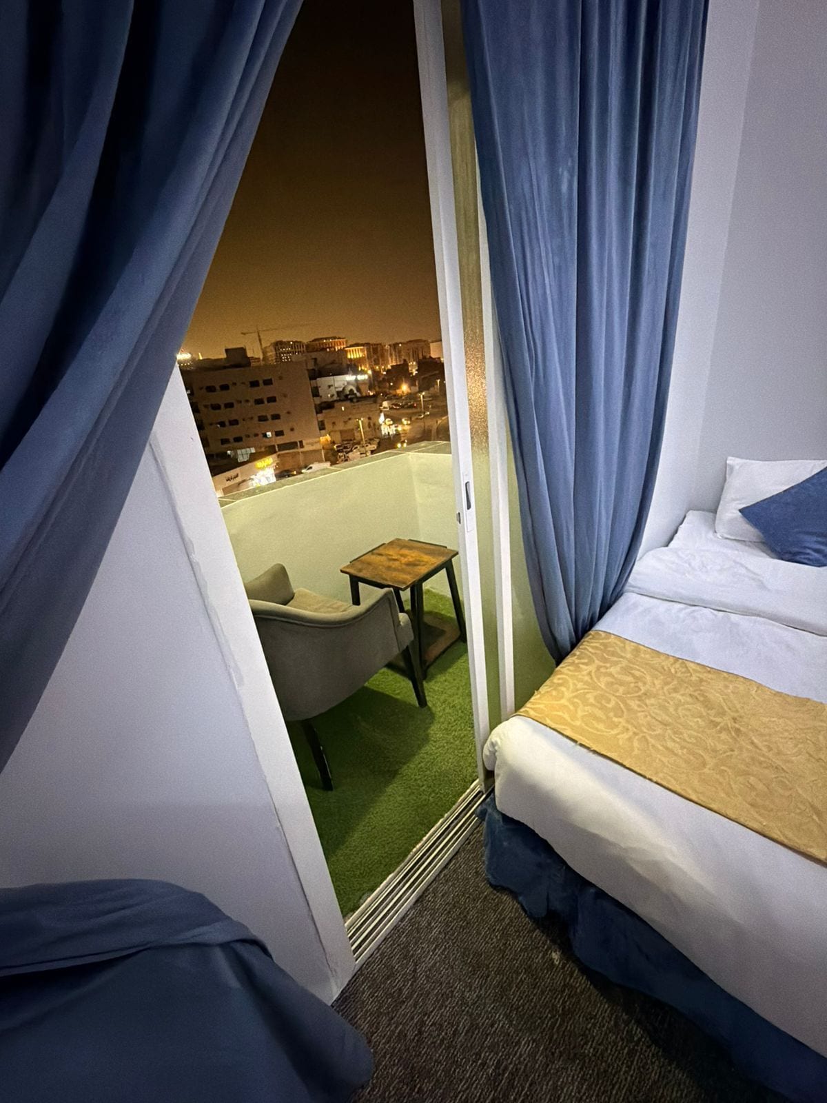 Habitación de hotel con cama, una silla, una mesita y vista a la ciudad de noche.