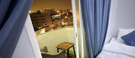 A hotel room with a bed, a chair, a small table, and a view of the cityscape at night.