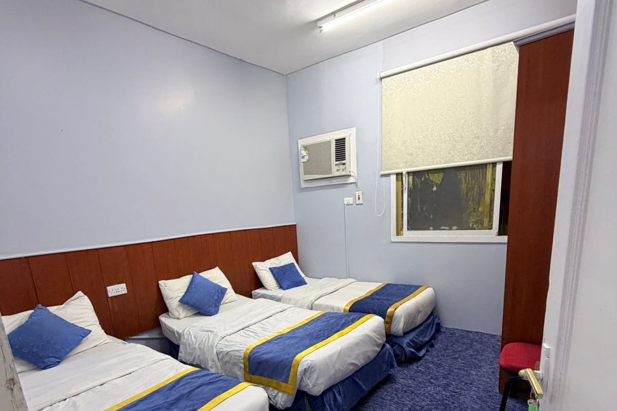 Habitación de hotel con tres camas, ventana con persianas, aire acondicionado y piso alfombrado.