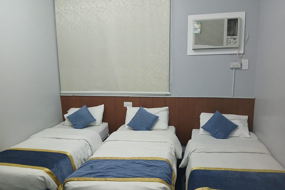 Habitación de hotel con tres camas individuales, cada una con almohada azul y colcha blanco y azul. Se observa un equipo de aire acondicionado instalado en la pared sobre las camas.