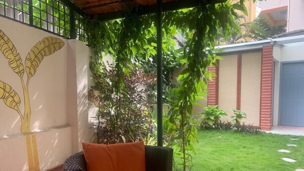 A patio area with a wicker sofa and cushions, a wall with a banana plant mural, and a well-maintained lawn. - Brham Store and Residency Brunton Road  (Bengaluru)