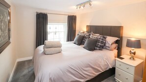 Een net opgemaakt bed met kussens en een opgerolde handdoek, een bedlampje en een raam met gordijnen.