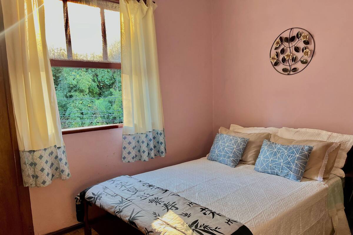 Quarto com cama, almofadas, janela com cortinas e um objeto decorativo na parede.