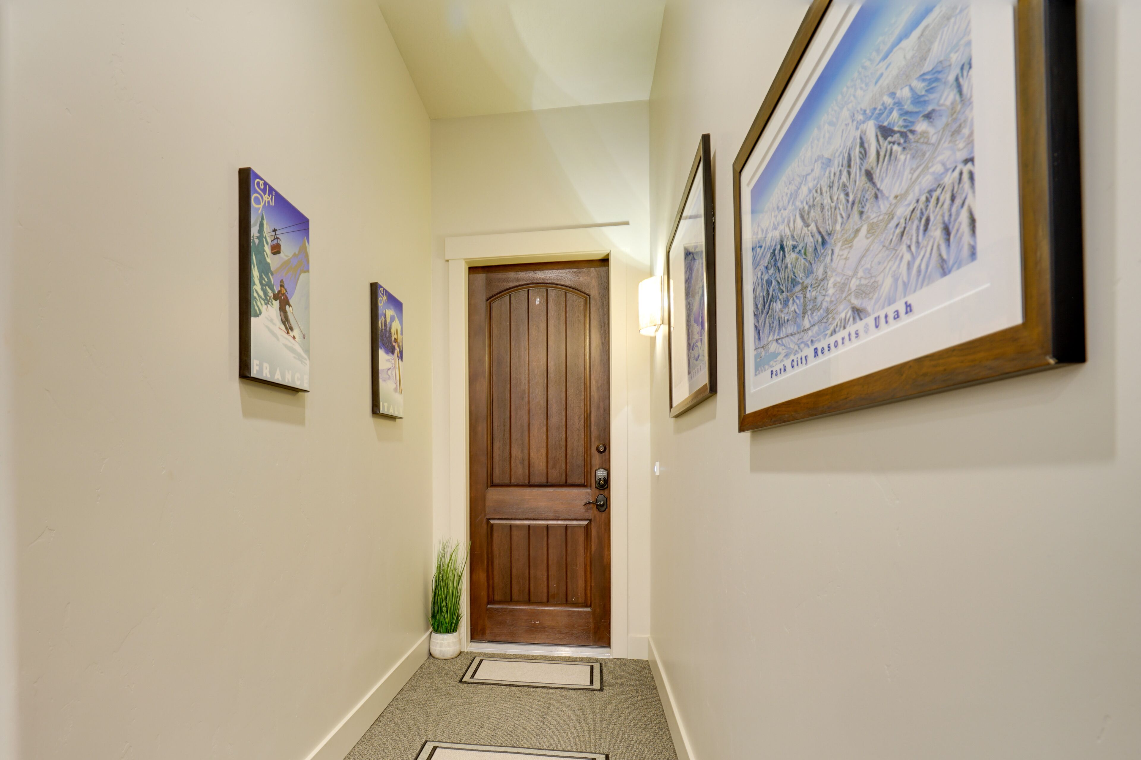 A hallway with a wooden door, framed pictures on the walls, and a small potted plant.