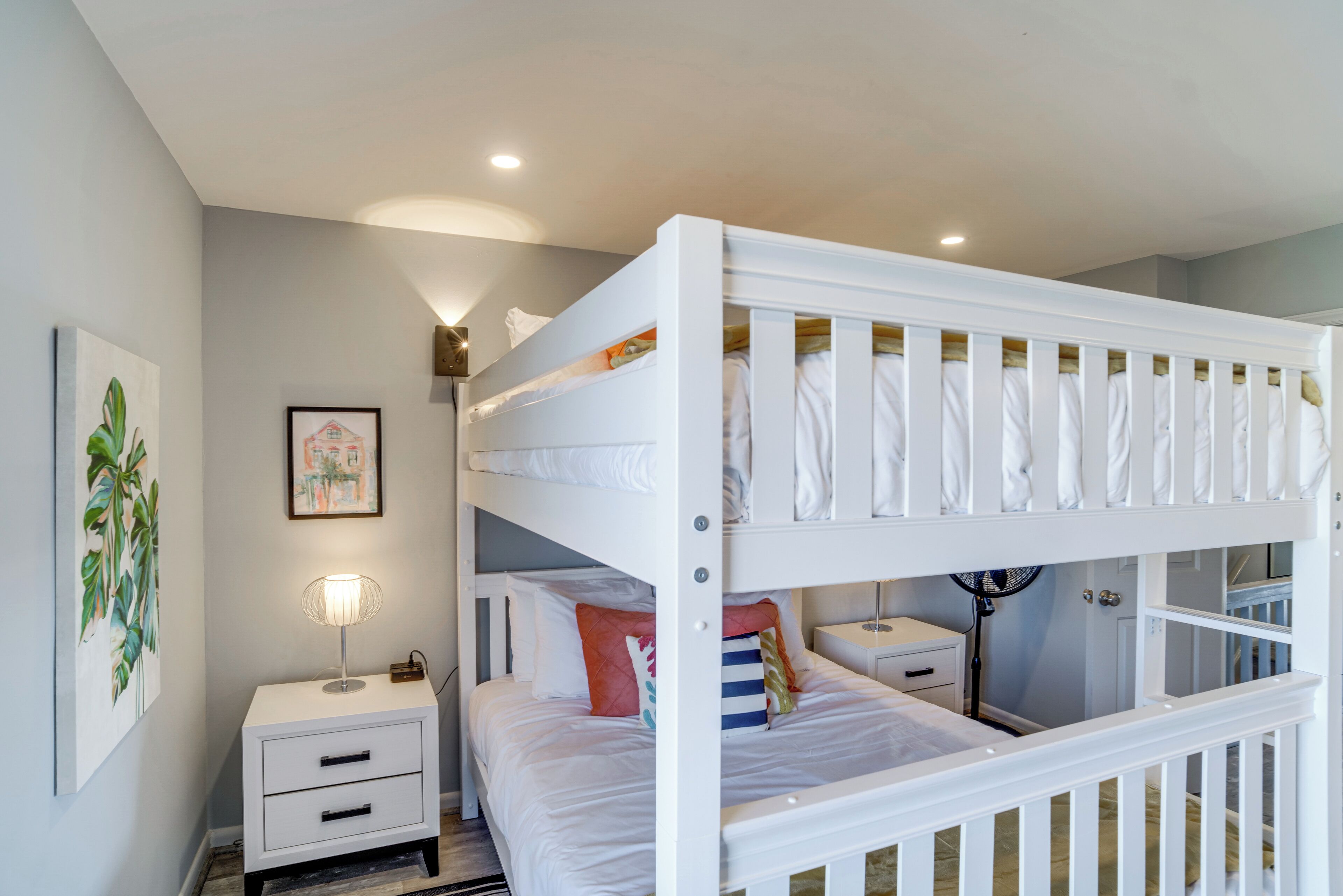 A bunk bed room with a white finish, a bedside table with a lamp, and a framed picture on the wall.