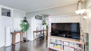 A living room with a large flat-screen TV, wooden flooring, white cabinets, and a bookshelf.