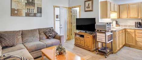 A living room with a sofa, coffee table, and a flat-screen TV. There is a kitchen area with wooden cabinets and a microwave.