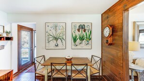 Een eethoek met een houten tafel en stoelen, een houten klok aan de muur en botanische prints erboven.