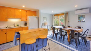 A modern kitchen with wooden cabinets, a central island with stools, a dining table with chairs, and a window with curtains. - Comfortable Lincoln Home: Walk to Main Street! (Lincoln)