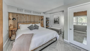 Une chambre avec un lit, une tête de lit dotée de lattes verticales, un panneau mural indiquant « SOYEZ NOTRE INVITÉ », une table de chevet avec une lampe et un banc près d’une porte coulissante.