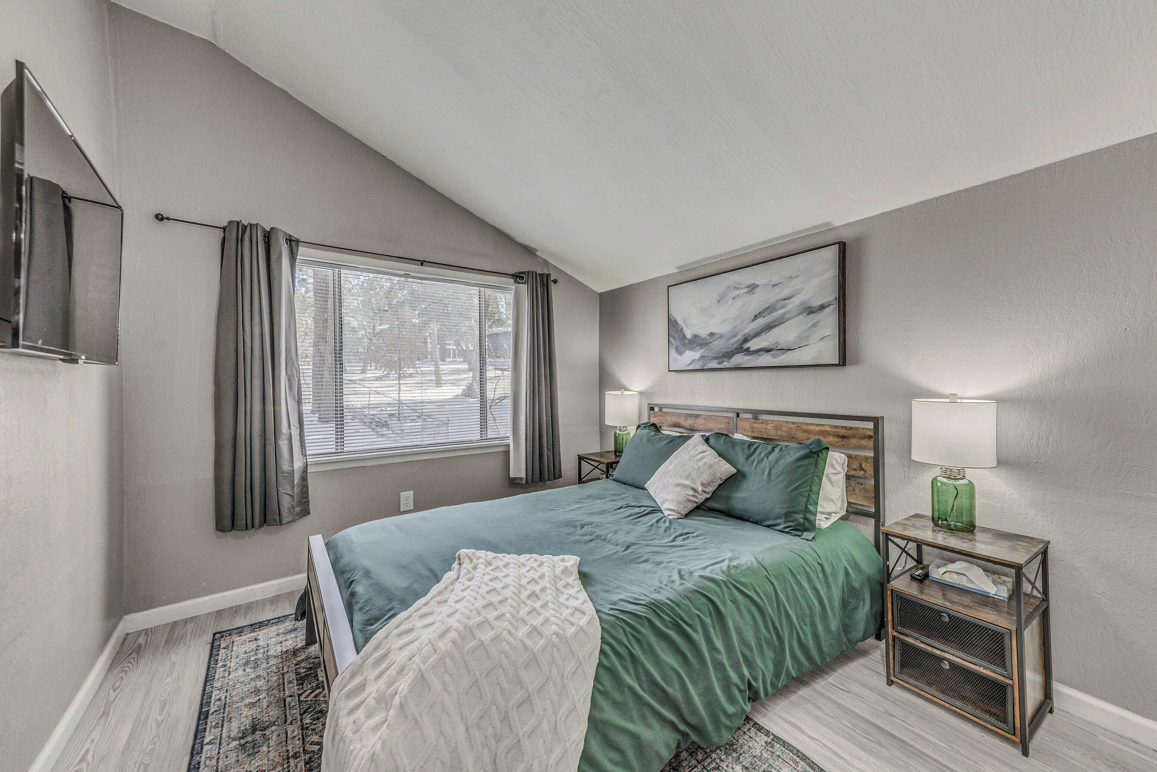 A bedroom with a bed, two bedside tables, a window with curtains, a wall-mounted TV, and a painting.