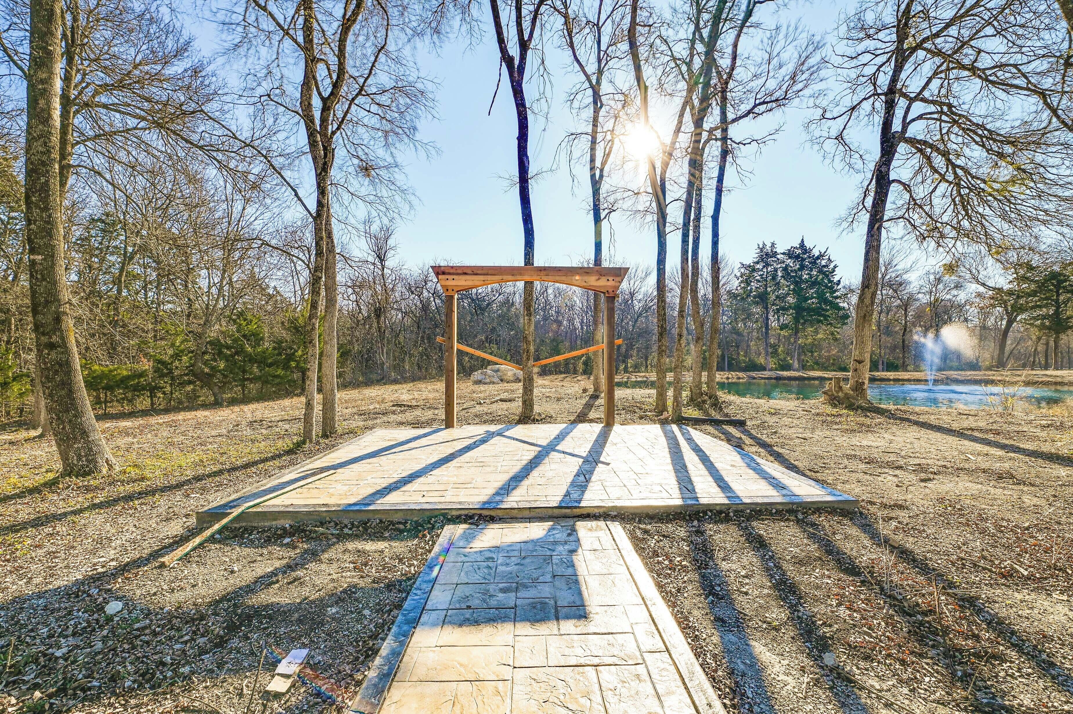 A wooden pergola structure in a park with a paved walkway leading to it.