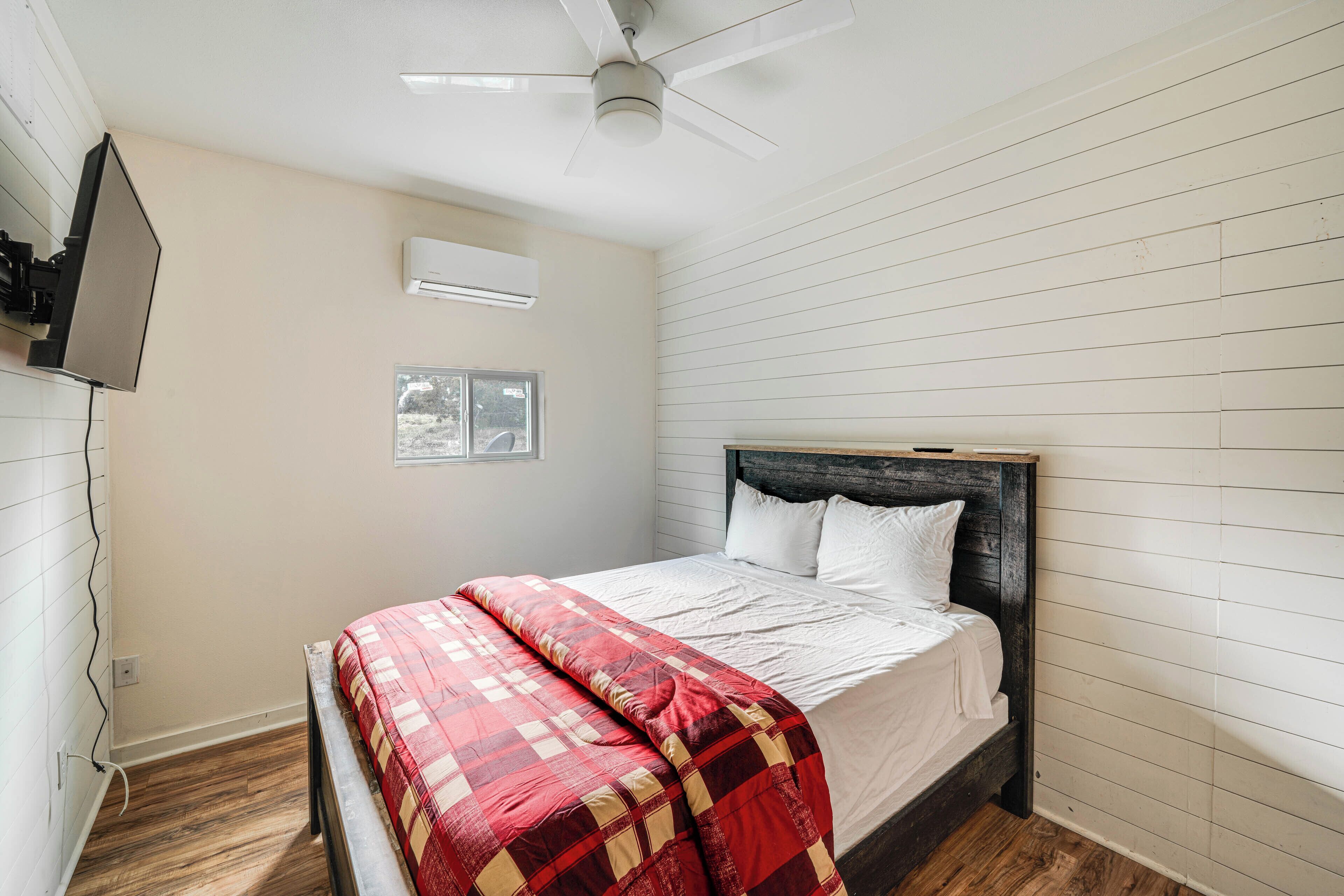 A bedroom with a bed, a ceiling fan, an air conditioner, a window, and wooden flooring.