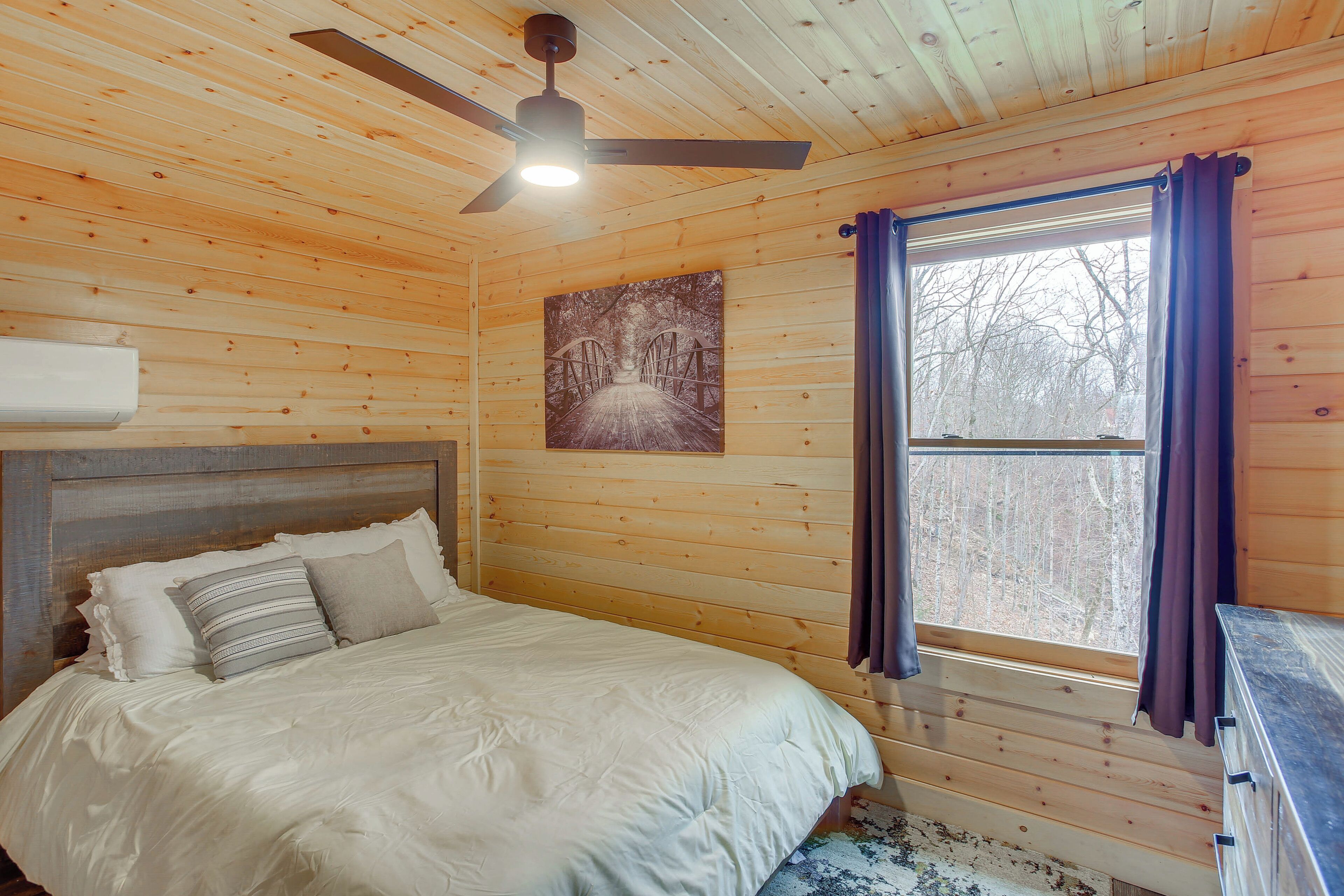 Un dormitorio de cabaña de madera con una cama, un ventilador de techo, una ventana con cortinas y un cuadro en la pared.