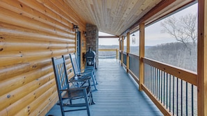 Une terrasse en bois avec une cheminée en pierre, des bancs et une vue sur un paysage boisé.
