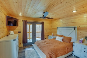 Quarto de cabana de madeira com cama, ventilador de teto, TV fixada na parede e porta deslizante para o exterior.