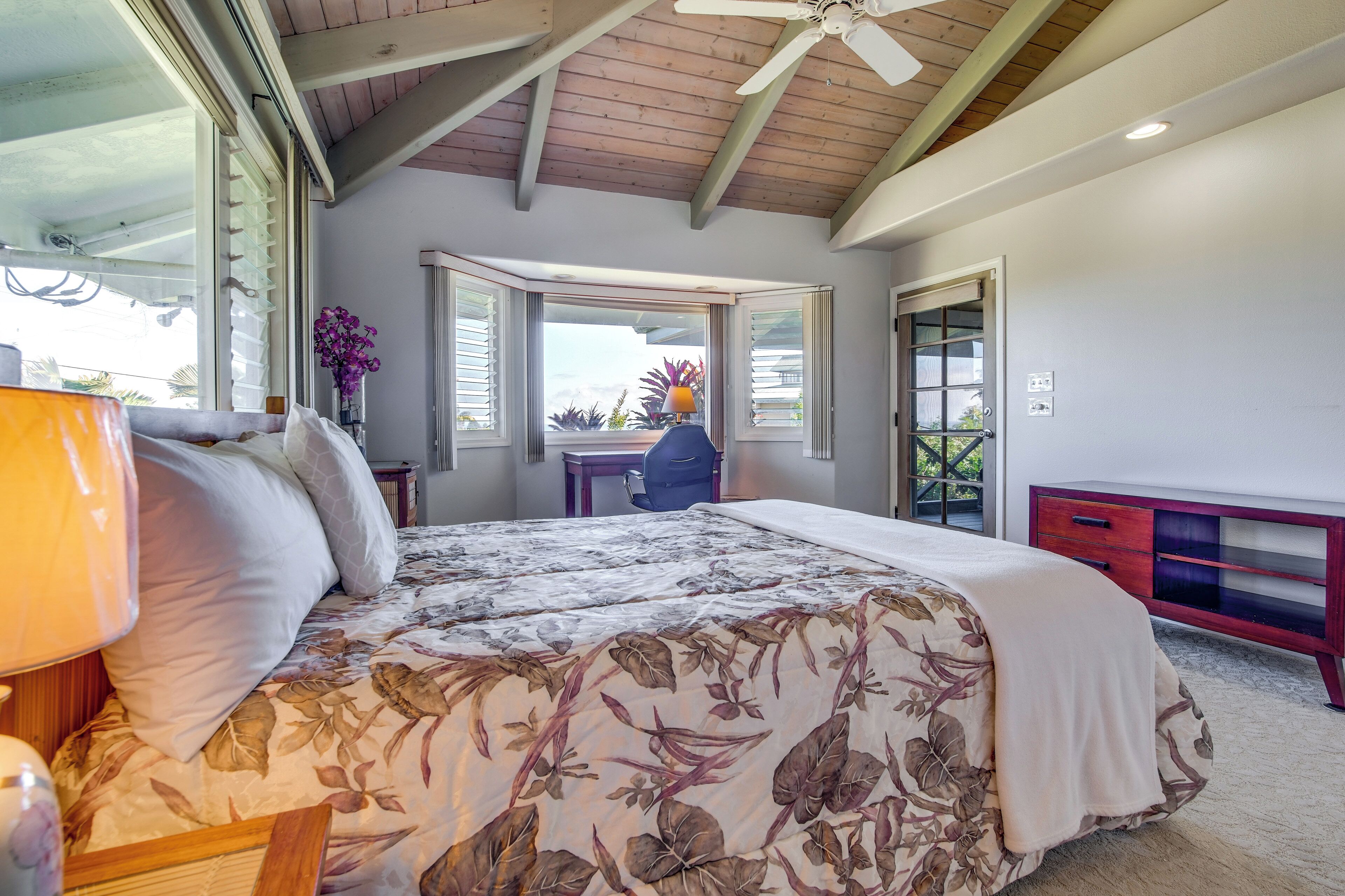 A bedroom with a bed, a desk, a chair, a window with a view, and a ceiling fan.