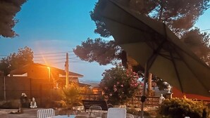 An outdoor patio with white metal chairs and a round table, surrounded by greenery and a large umbrella. - Vista al Mar, Piscina y Paz: tu Escape Perfecto en Esta Casa Rural Única (Sant Miquel)