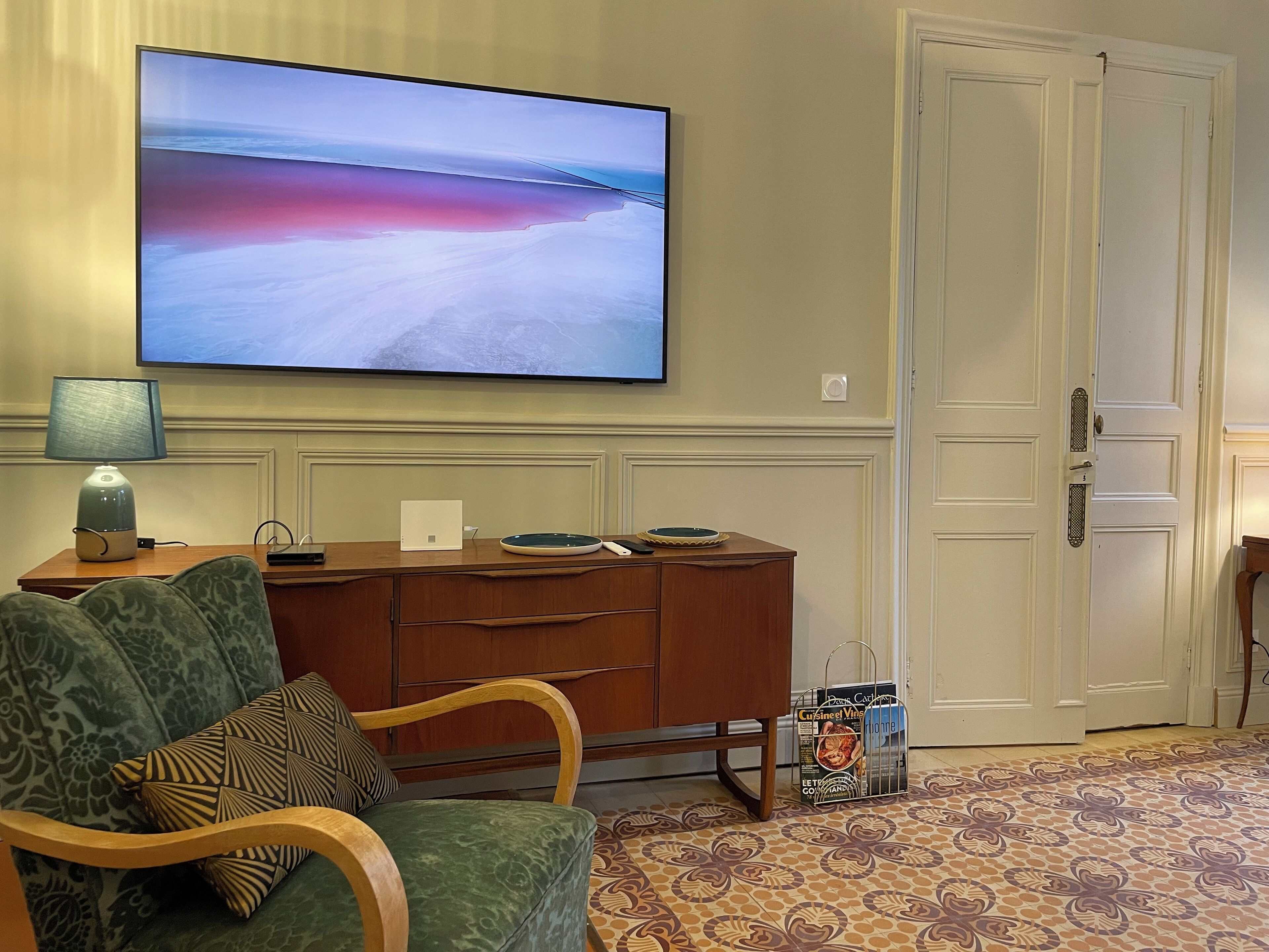 Ein Wohnzimmer mit einem an der Wand befestigten Flachbildfernseher, einem Holztisch mit Lampe, einem grünen Sofa mit gemusterten Kissen, einer Holzkabine und einem gemusterten Teppich.