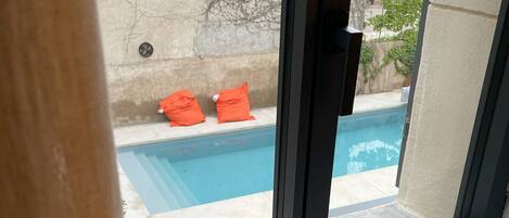 A view through a window of a swimming pool with two orange pillows on the edge, a concrete deck, and a building with a sign that reads "EPICERIE" in the background.