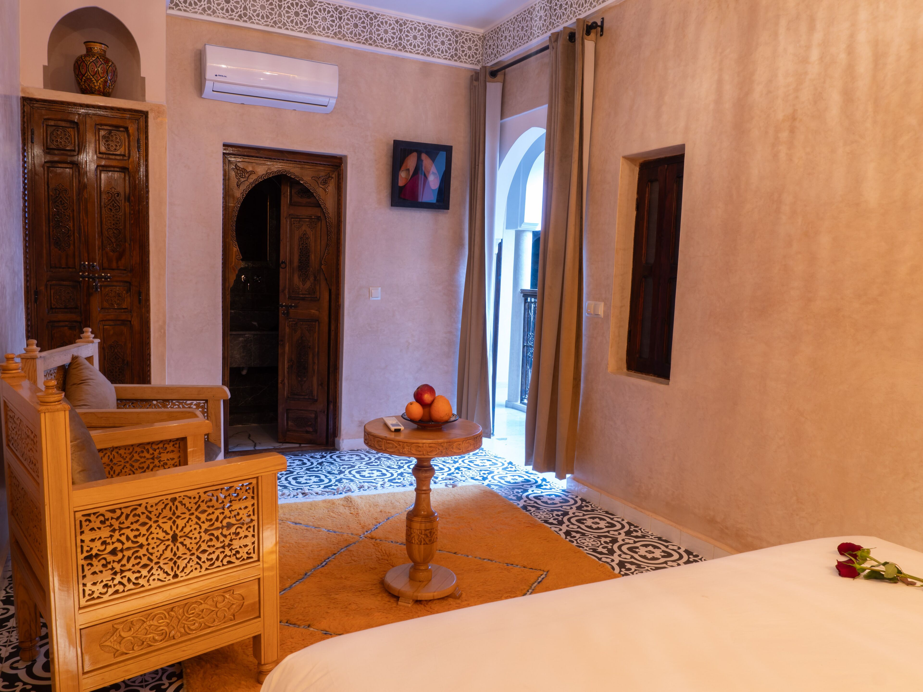 Una habitación con una cama, un banco de madera, una pequeña mesa con fruta, un florero y un cuadro.