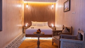 A room with a bed, two bedside tables with lamps, a small round table with a fruit bowl, a chair, and a decorative wall piece. - Click Riad & Spa (Marrakech)