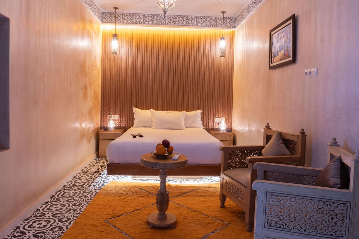 Una habitación con cama, dos mesitas de noche con lámparas, una pequeña mesa redonda con frutero, una silla y un adorno en la pared.