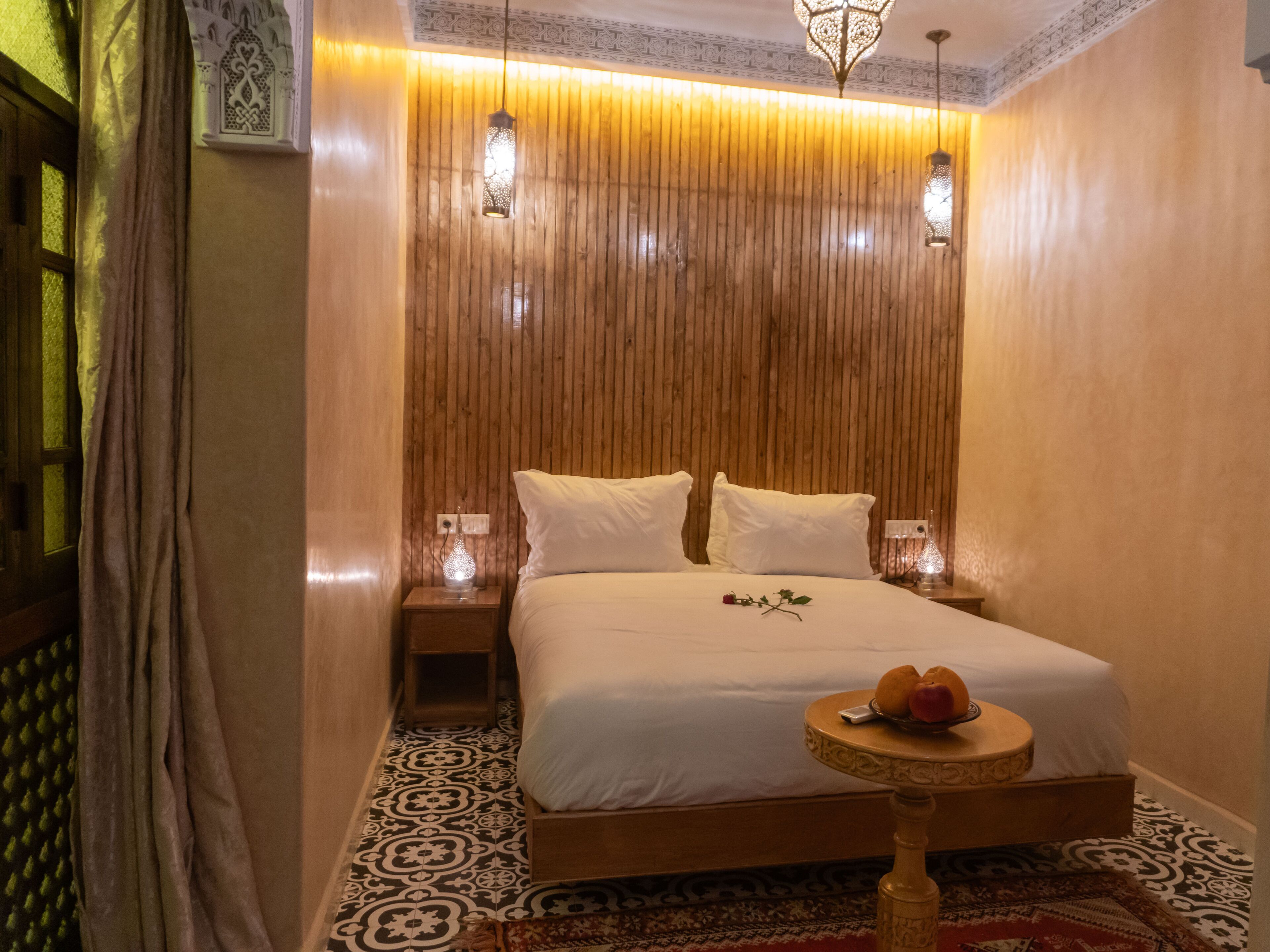 A bedroom with a wooden headboard, a bed with white linens, a bedside table with a lamp, and a small round table with fruits.