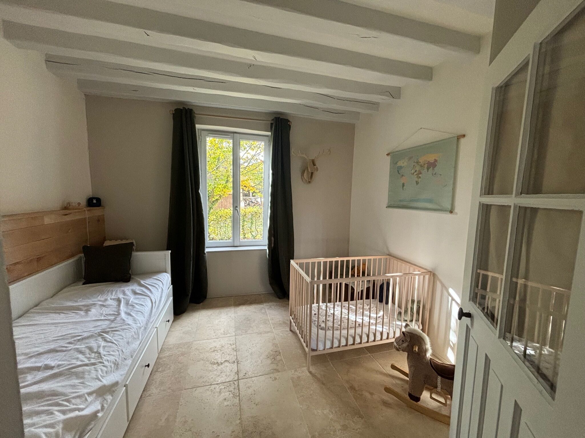 Una habitación con cama, cuna, un caballito de madera, ventana con cortinas, un mapa mundial en la pared y cabecera de madera.