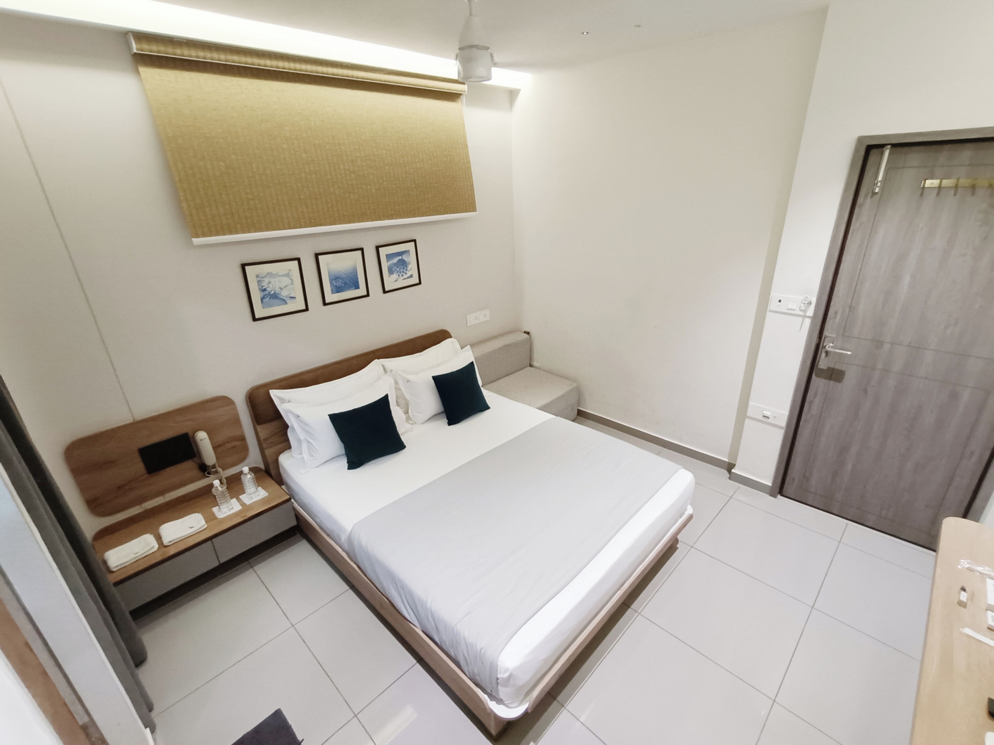 A hotel room with a bed, bedside tables, a nightstand, a wooden headboard, a door, and a wall-mounted light fixture.