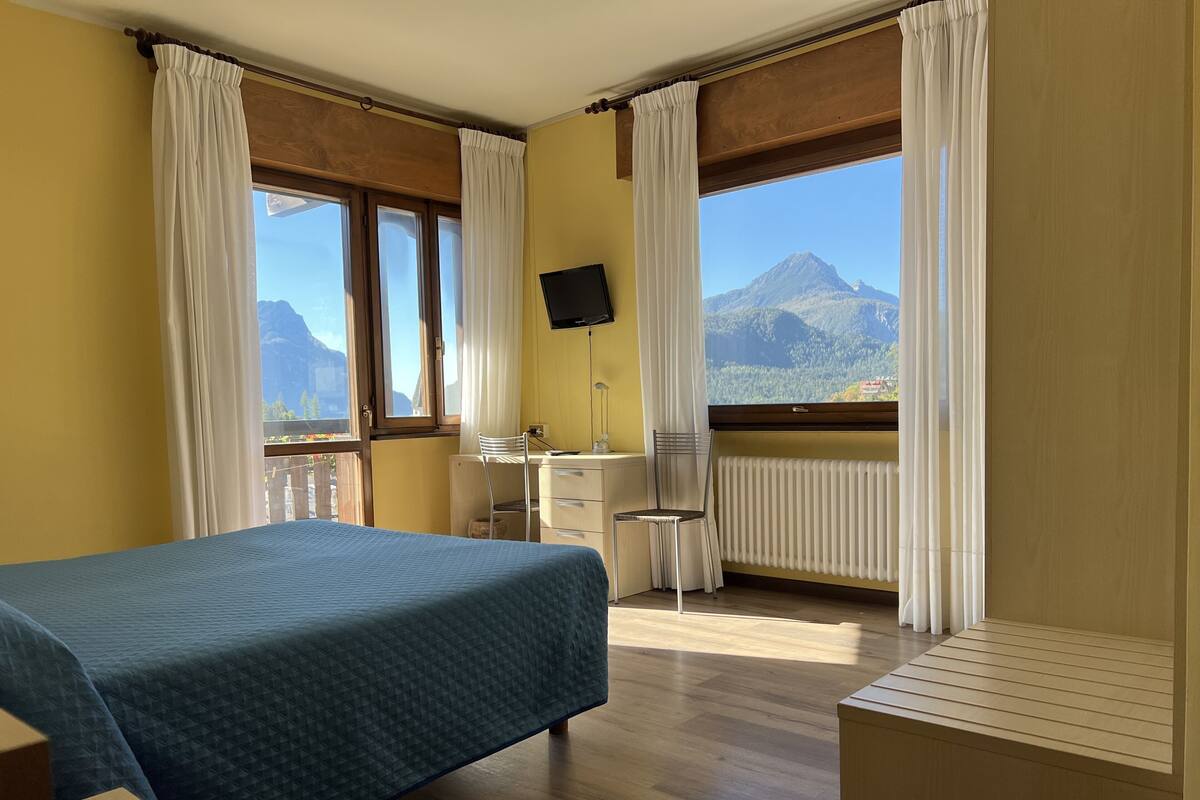 Habitación de hotel con cama, escritorio, silla y un amplio ventanal con vistas a la montaña.