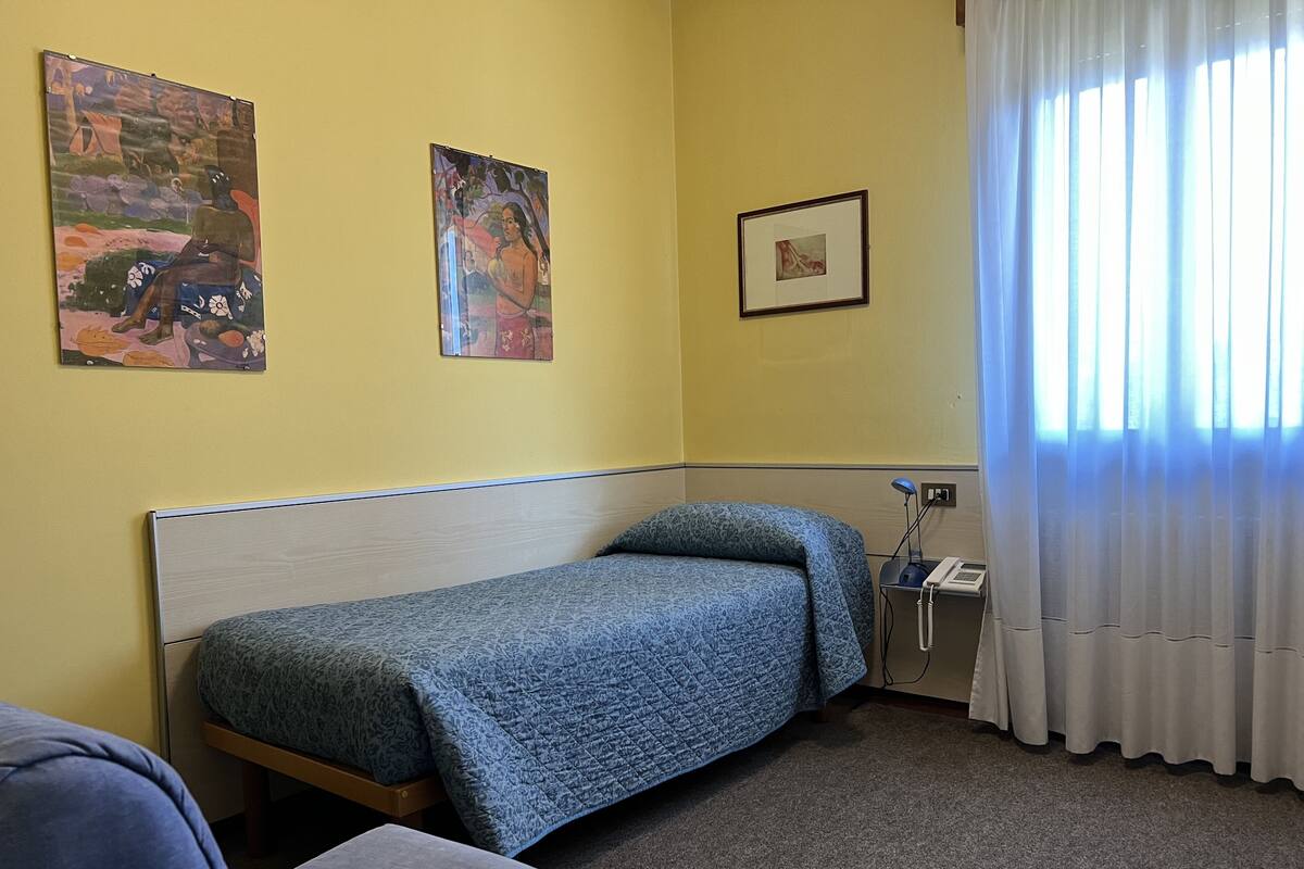 Una cama individual con una colcha azul, una mesita de noche con una lámpara, un teléfono y un cuadro en la pared.