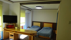 Une chambre d’hôtel avec un bureau en bois, un téléviseur à écran plat, deux lits avec une literie bleue, un luminaire au plafond et une fenêtre avec des stores.