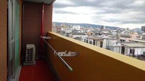 A balcony with a red floor, a metal handrail, and a view of a cityscape with buildings and mountains in the background. - It has a kitchen, closet, shoe closet, washing machine, balcony and parking space. Clothes can be dried on the balcony, or room-dried. The 46 sq. m. of space is ideal for long-term stays. (大阪市)
