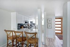A modern kitchen with white cabinets, a central island, and a dining area with wicker chairs. - The Designer Condo, Sleek | Centrally Located! (Savannah)